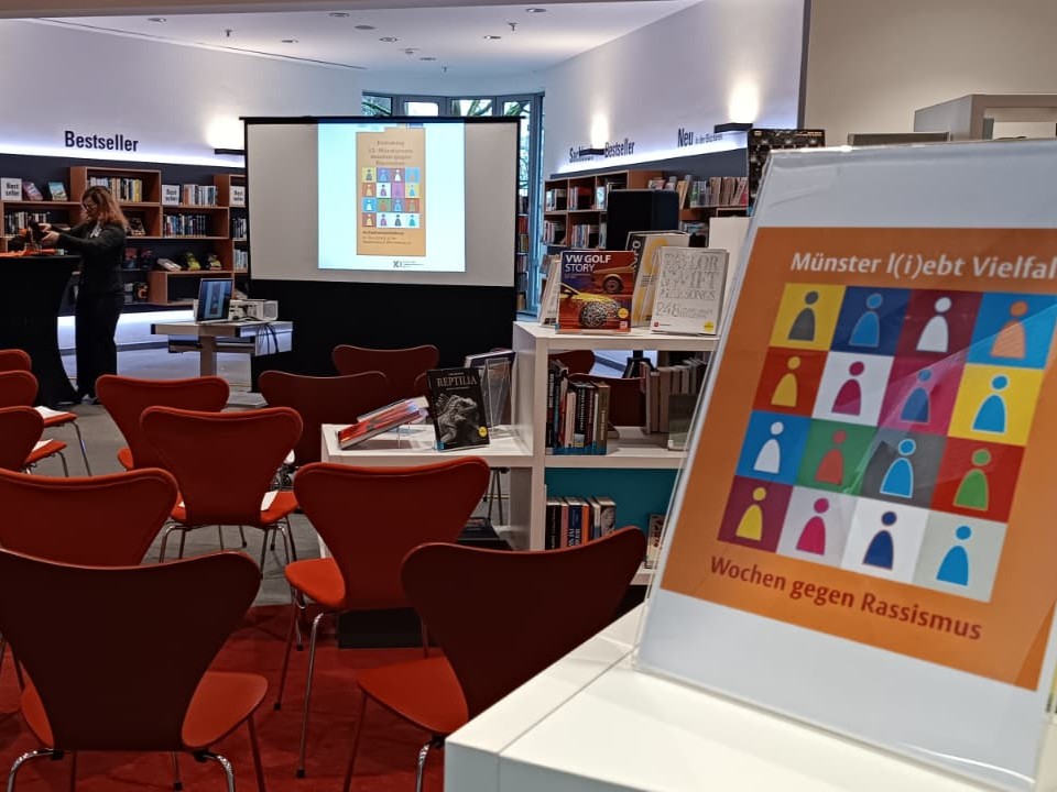 Im Vordergrund ein Schild der Wochen gegen Rassismus, dahinter mehrere rote Stühle, davor eine Leinwand und mehrere Stehtische