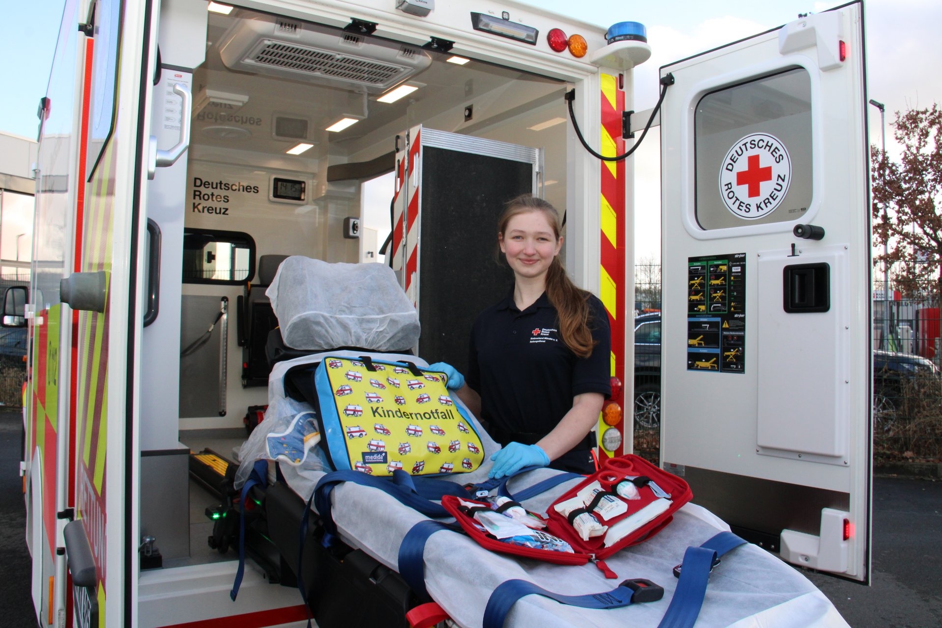 Eine junge Frau steht neben der ausgefahrenen Trage eines Krankentransportwagens des DRK. Auf der Liege befindet sich Material.