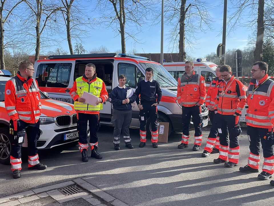 Eine Gruppe von Einsatzkräften in Einsatzkleidung stehen im Halbkreis. Eine Person mit zusätzlicher gelber Weste und Klemmbrett spricht zu ihnen.