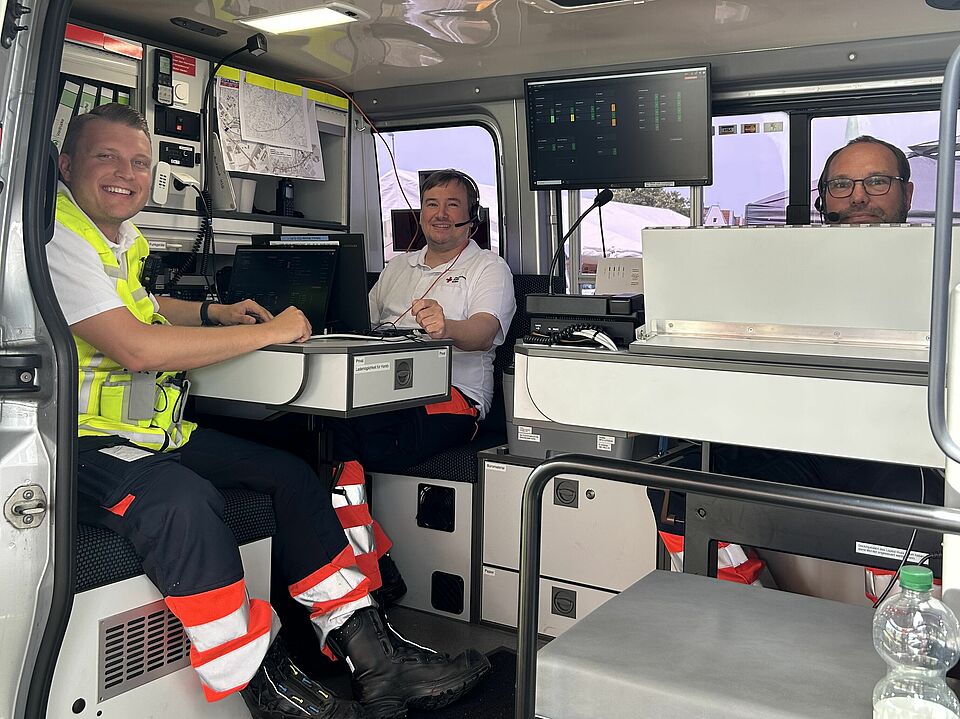 Drei Einsatzkräfte sitzen lächelnd im Einsatzleitwagen mit Headsets und Monitoren, bereit zur Koordination von Rettungseinsätzen.