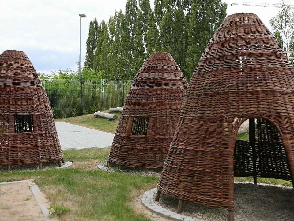 Mehrere kegelartige Hütten aus geflochtenen Weidenruten auf einer Wiese, umgeben von Bäumen und einem Zaun unter leicht bewölktem Himmel.