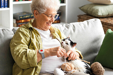 Eine Seniorin sitzt mit einer Katze auf dem Sofa und strickt. Dabei trägt sie einen roten Hausnotruf-Sender am Handgelenk.
