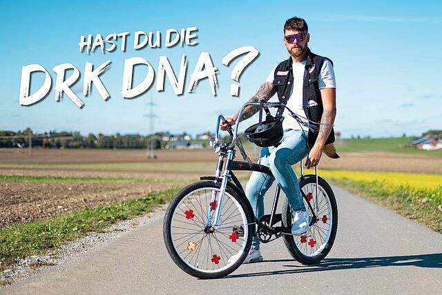 Eine Person fährt mit dem Fahrrad auf einer Landstraße umgeben von Feldern und einigen Gebäuden im Hintergrund. In den Speichen befinden sich Rotkreuzreflektoren. Text im Bild: „Hast du die DRK DNA?“