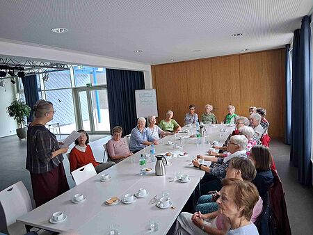 Eine Gruppe von älteren Leuten sitzt um einen großen Tisch, auf dem Getränke und Geschirr stehen, herum. Im Vordergrund steht eine Fraz und spricht zur Gruppe.