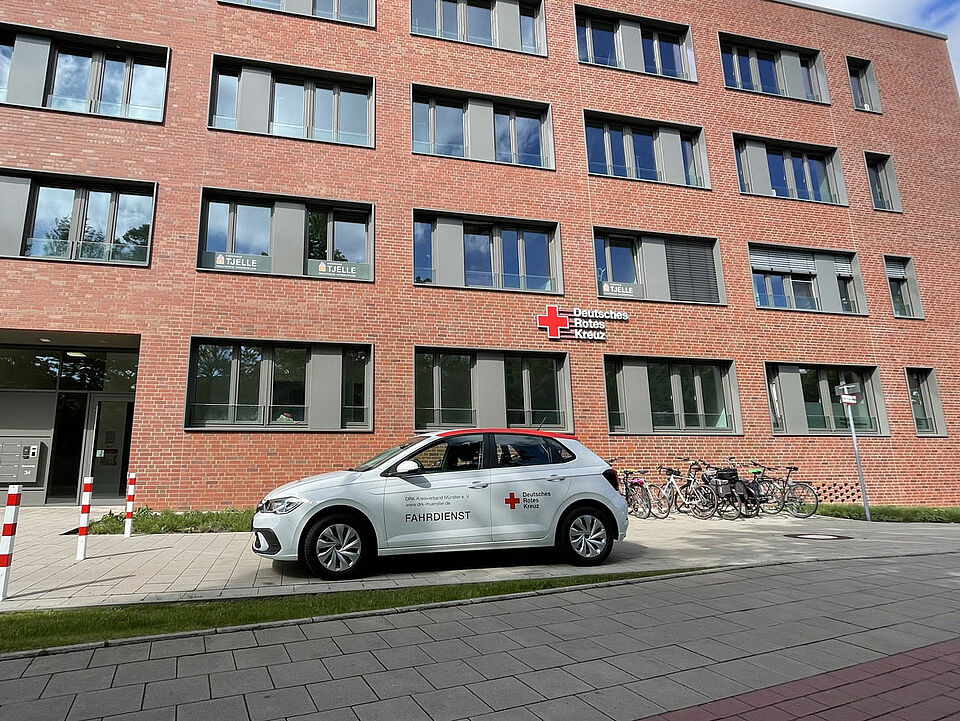 Modernes Backsteingebäude mit großem DRK-Schild. Vor dem Gebäude steht ein weißer Kleinwagen mit DRK-Logo und der Beschriftung "Fahrdienst"