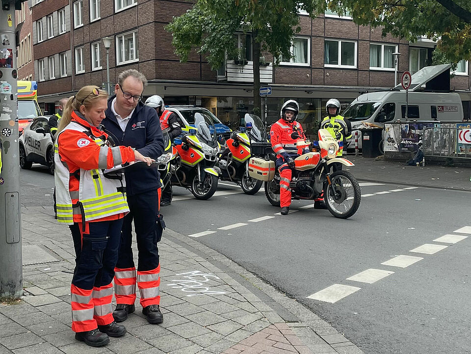 Zwei Einsatzkräfte besprechen sich neben einer Straße, auf der Motorräder in Einsatzkleidung warten