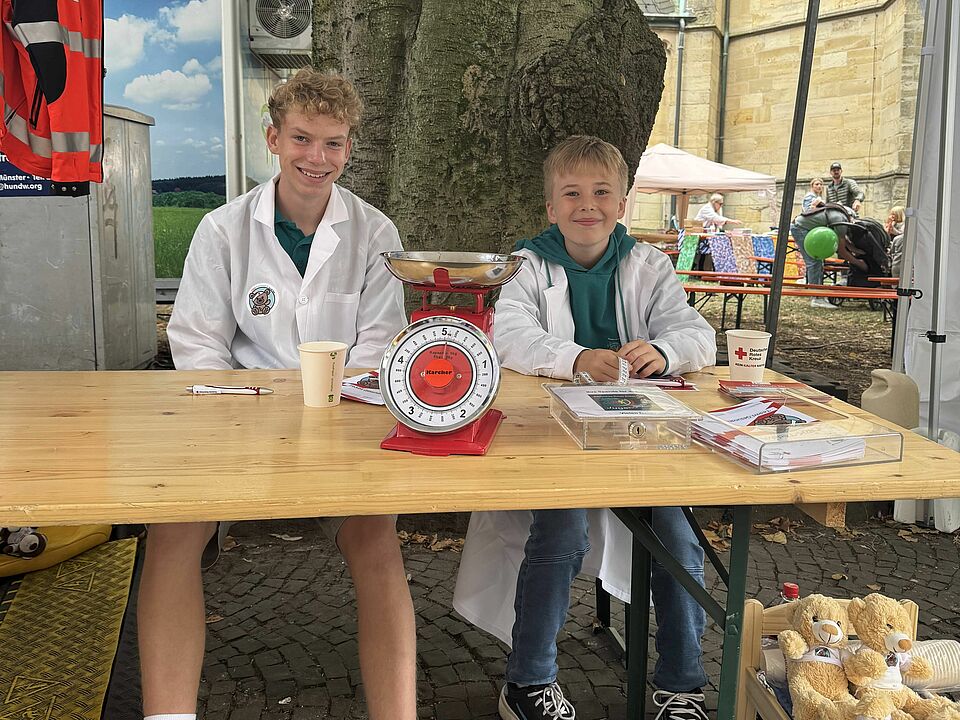 Zwei Kinder sitzen an einem Tisch unter einem Zelt. Auf dem Tisch steht eine Waage. Die Kinder tragen türkise T-Shirts vom Jugendrotkreuz und einen weißen Kittel.