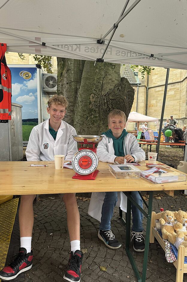 Zwei Kinder sitzen an einem Tisch unter einem Zelt. Auf dem Tisch steht eine Waage. Die Kinder tragen türkise T-Shirts vom Jugendrotkreuz und einen weißen Kittel.