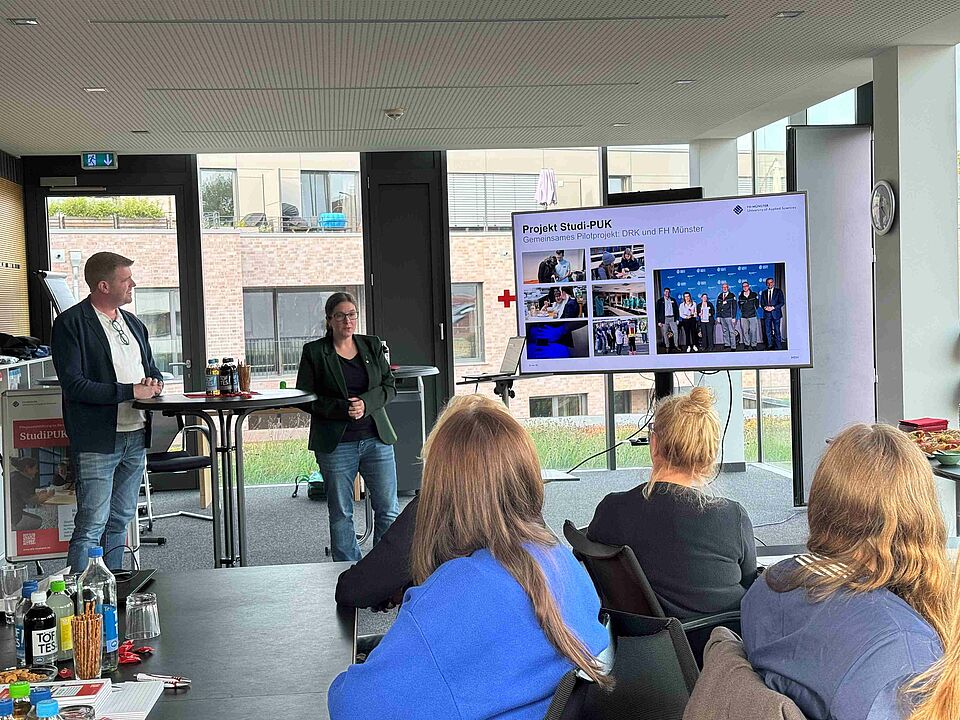 Zwei Personen halten eine Präsentation, die auf einem Bildschirm zu sehen ist, zum Thema "StudiPUK" vor einem Publikum