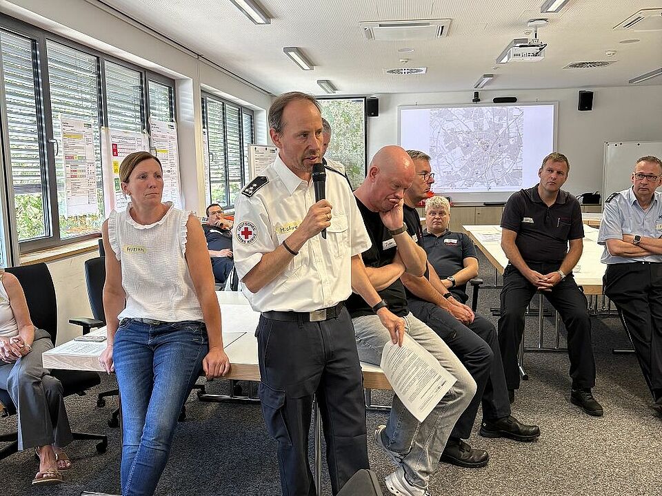 Planübung MANV Mehrere Personen stehen in einem Halbkreis in einem Raum und hören einer Person mit Mikrofon zu. Im Hintergrund sind eine Lagekarte und Plakate zu sehen.