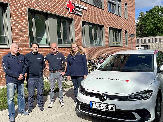 Vier Personen stehen neben einem DRK-Auto vor einem modernen Backsteingebäude, an dem ein DRK-Logo zu sehen ist