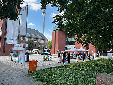 Vorplatz eines modernen Gebäudes mit mehreren Pavillions, Tischen und Stühlen, einem Kletterberg, großen DRK-Fahnen und mehreren Personen