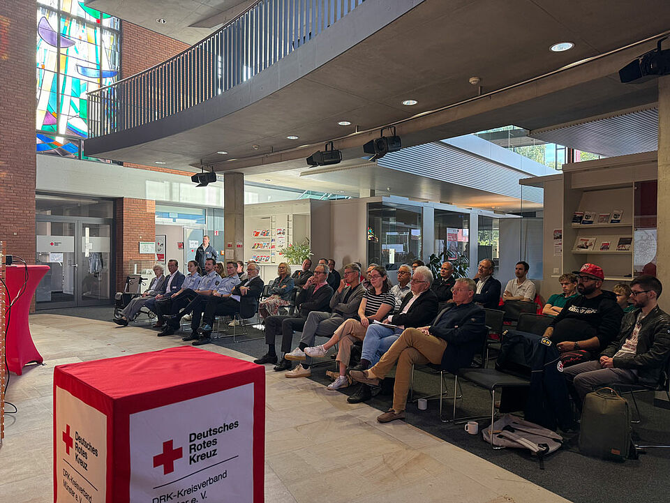 Ein Publikum von etwas 30 Personen in einem modernen Gebäude, welches auf eine Bühne schaut, von vorne fotografiert.
