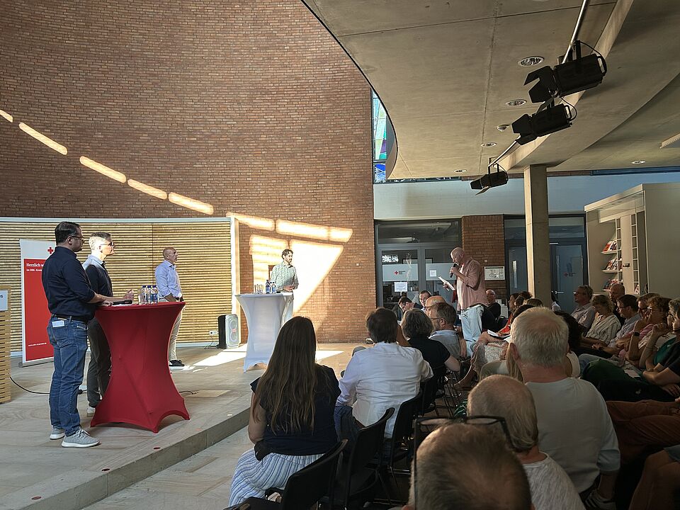 Vier Personen stehen bei einer Veranstaltung vor einem sitzenden Publikum. Zwei stehen hinter einem roten Tisch, zwei an einem weißen Rednerpult. Eine Person im Publikum steht und hält ein Mikrofon in der Hand. Der Raum hat Backsteinwände und große Fenster.
