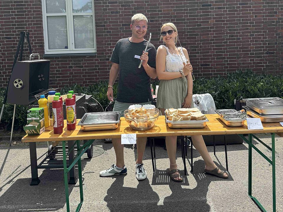 Zwei Personen stehen hinter einem Tisch mit Grillutensilien und hinter ihnen stehen zwei Grills