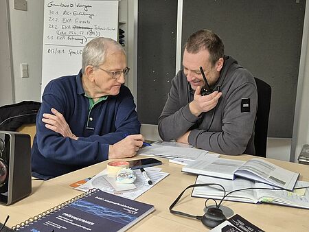Zwei Personen sitzen an einem Tisch, vor ihnen liegen Bücher und weitere Unterlagen. Eine Person hält ein Funkgerät in der Hand.