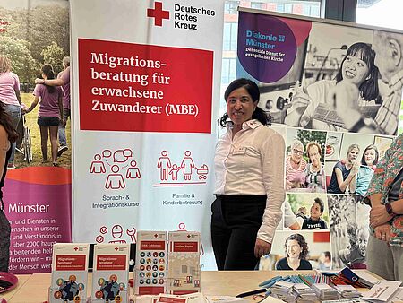 Eine Frau steht hinter einem Tisch mit Flyern und weiteren Materialien und neben einem Roll-Up mit dem DRK-Logo und "Migrationsberatung für erwachsene Zugewanderte"
