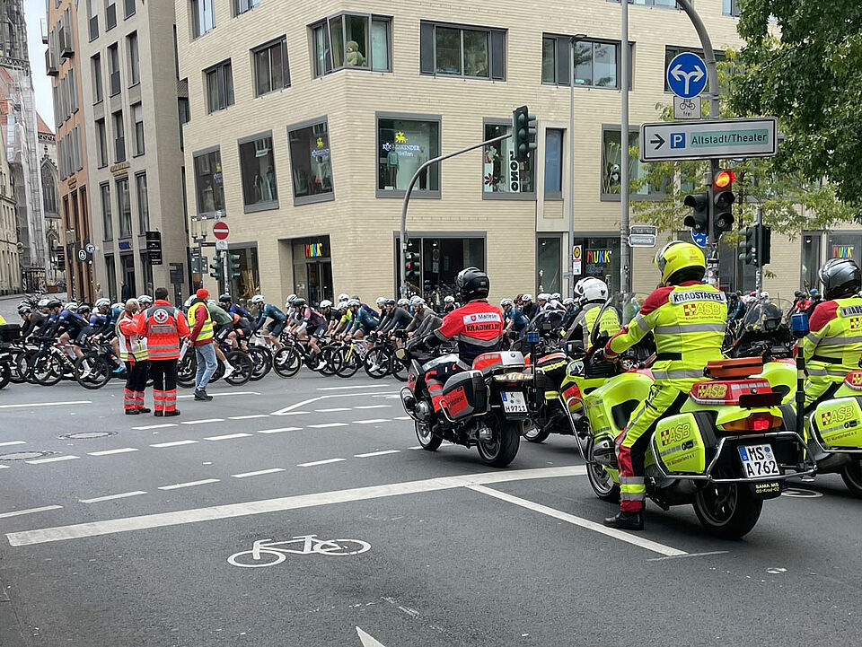 Rettungskräfte auf Motorrädern stehen in Position, auf der Straße ist ein Feld von Radfahrer*innen. Zwei Einsatzkräfte koordinieren die Situation. Im Hintergrund ist ein Kirchturm zu sehen.