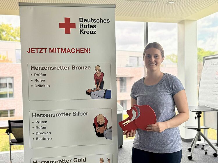 In einem hellen Schulungsraum steht eine junge Frau und hält ein Schluckmodell n in der Hand. Neben ihr steht ein Roll-Up des Deutschen Roten Kreuzes mit dem Titel „Jetzt mitmachen“ und Informationen zu den drei Stufen des „Herzensretter“-Programms: Bronze, Silber und Gold