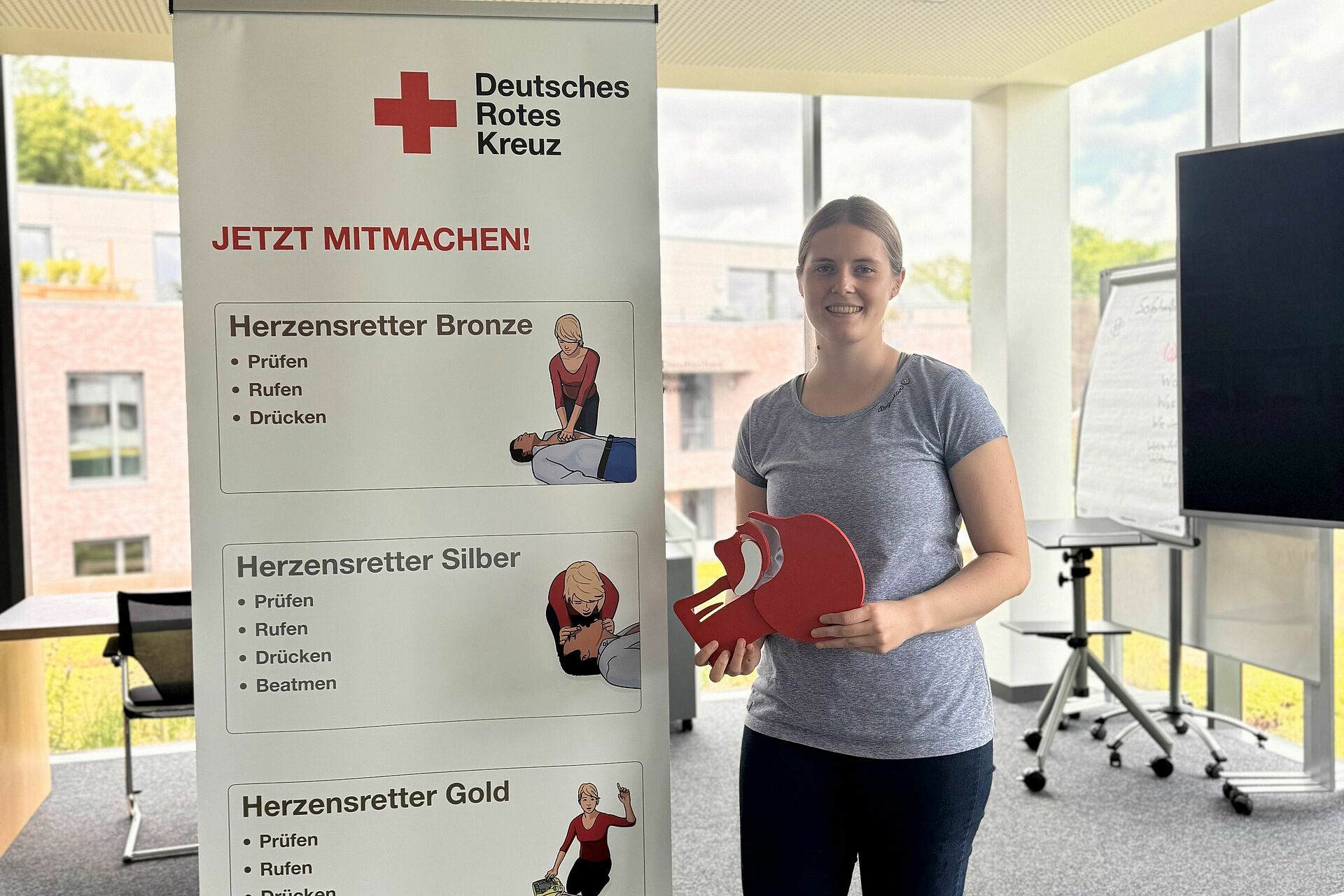 In einem hellen Schulungsraum steht eine junge Frau und hält ein Schluckmodell n in der Hand. Neben ihr steht ein Roll-Up des Deutschen Roten Kreuzes mit dem Titel „Jetzt mitmachen“ und Informationen zu den drei Stufen des „Herzensretter“-Programms: Bronze, Silber und Gold