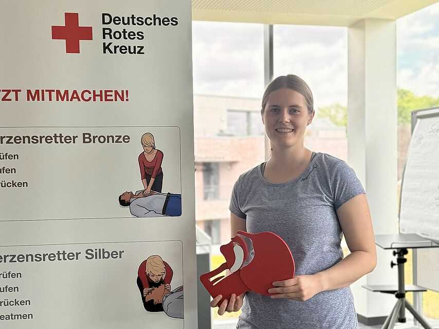 In einem hellen Schulungsraum steht eine junge Frau und hält ein Schluckmodell n in der Hand. Neben ihr steht ein Roll-Up des Deutschen Roten Kreuzes mit dem Titel „Jetzt mitmachen“ und Informationen zu den drei Stufen des „Herzensretter“-Programms: Bronze, Silber und Gold