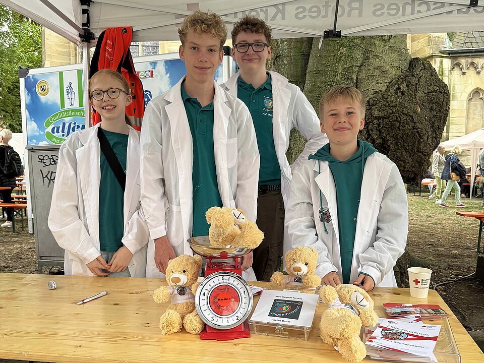 Gruppenfoto vier Kinder und Jugendlichen, die unter einem weißen Pavillion hinter einem Tisch stehen. Sie tragen türkise JRK-Oberteile und einen weißen Kittel. Auf dem Tisch steht eine Waage und mehrere Teddys sowie Flyer.