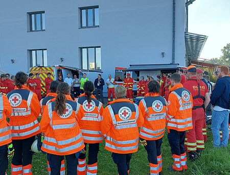 Personen in Einsatzkleidung stehen im Kreis, auf der einen Seite stehen Personen mit zusätzlichen Westen, die Unterlagen in der Hand halten, Einsatzleitungswägen von DRK und DLRG stehen im Hintergrund