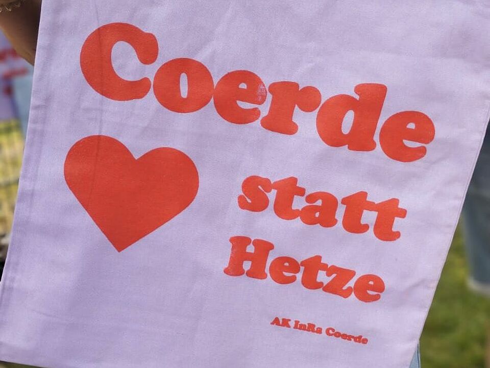 Eine Person hält eine hellviolette Stofftasche in der Hand. Auf der Tasche steht in roter Schrift: „Coerde - Herz statt Hetze“ und darunter „AK InRa Coerde“