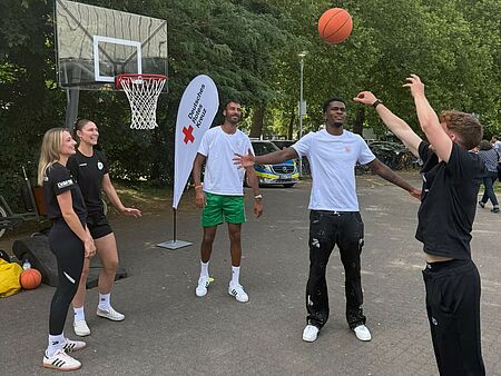 Fünf junge Menschen stehen vor einem Basketballkorb und werfen einen Basketball. Im Hintergrund ist eine DRK-Beachflag zu sehen.
