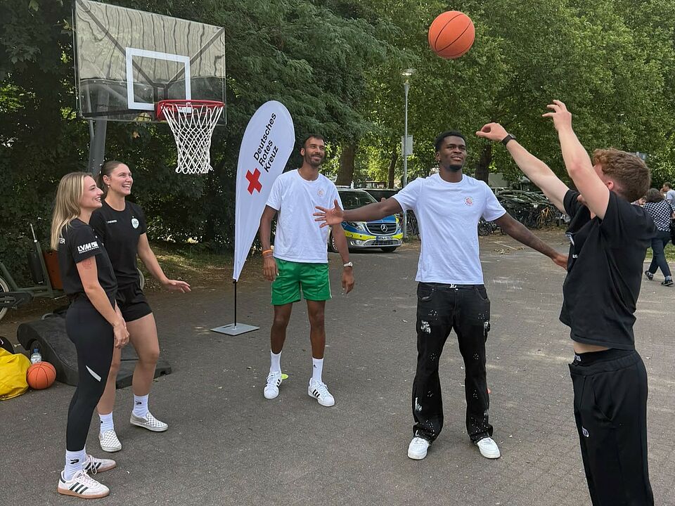 Fit in Berg Fidel Fünf junge Menschen stehen vor einem Basketballkorb und werfen einen Basketball. Im Hintergrund ist eine DRK-Beachflag zu sehen.