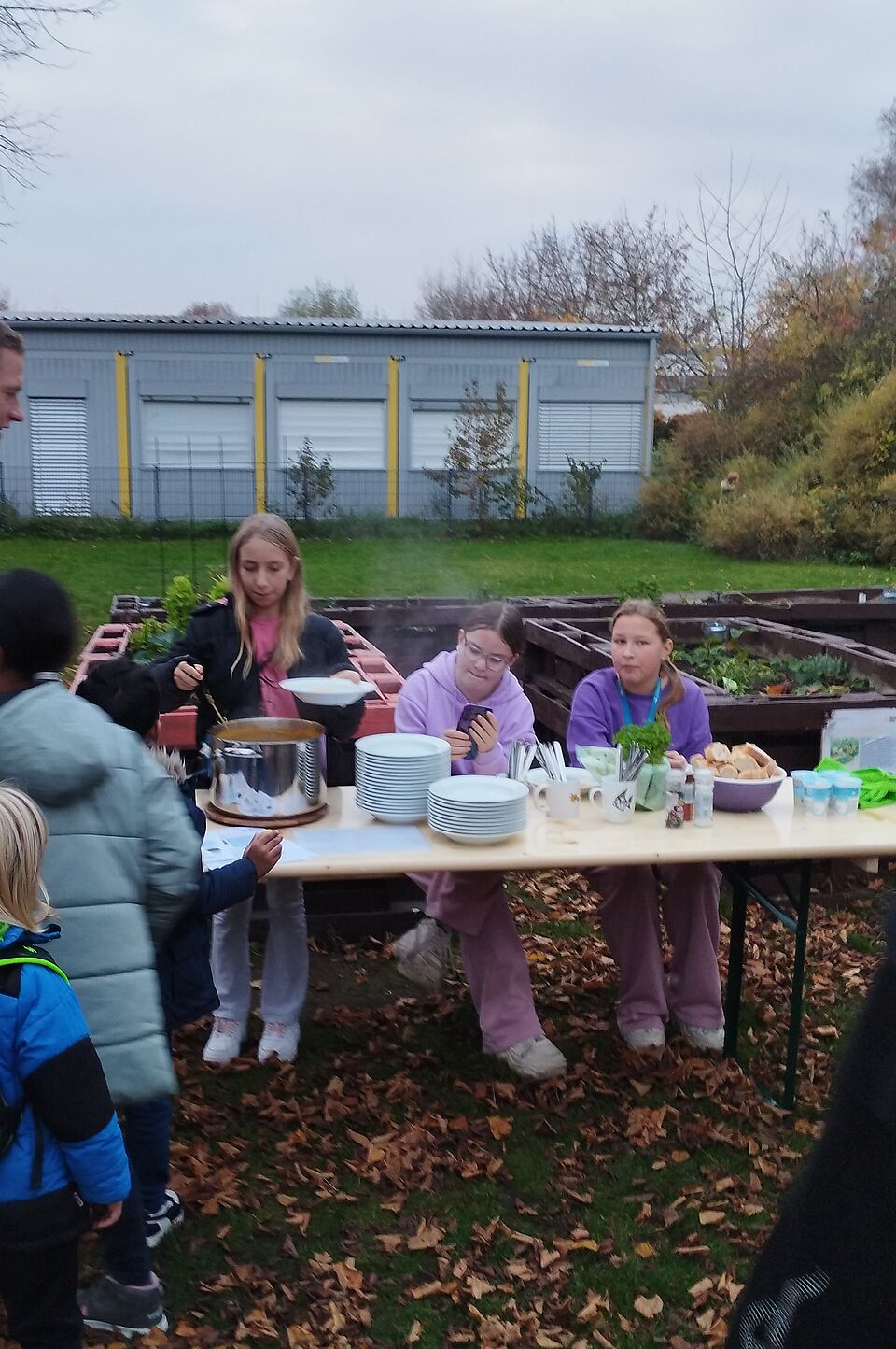 Auf dem Bild ist eine kleine Outdoor-Veranstaltung zu sehen. Drei Mädchen stehen an einem Tisch, der mit Tellern, Bechern und anderen Utensilien gedeckt ist. Eine der Mädchen serviert eine warme Mahlzeit aus einem großen Topf. Einige Kinder, die in winterlicher Kleidung wie Jacken und Mützen gekleidet sind, stehen in der Nähe des Tisches und warten darauf, ihr Essen zu bekommen. Der Hintergrund zeigt einen Gartenbereich mit Hochbeeten und bunten Herbstblättern, die den Boden bedecken