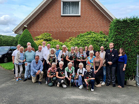 Eine Gruppe von Personen steht vor einer grünen Hecke, im Hintergrund ist ein Haus zu sehen.