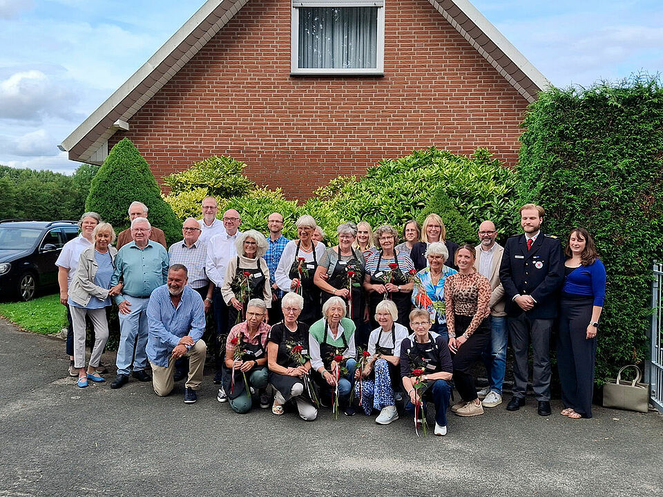Eine Gruppe von Personen steht vor einer grünen Hecke, im Hintergrund ist ein Haus zu sehen.