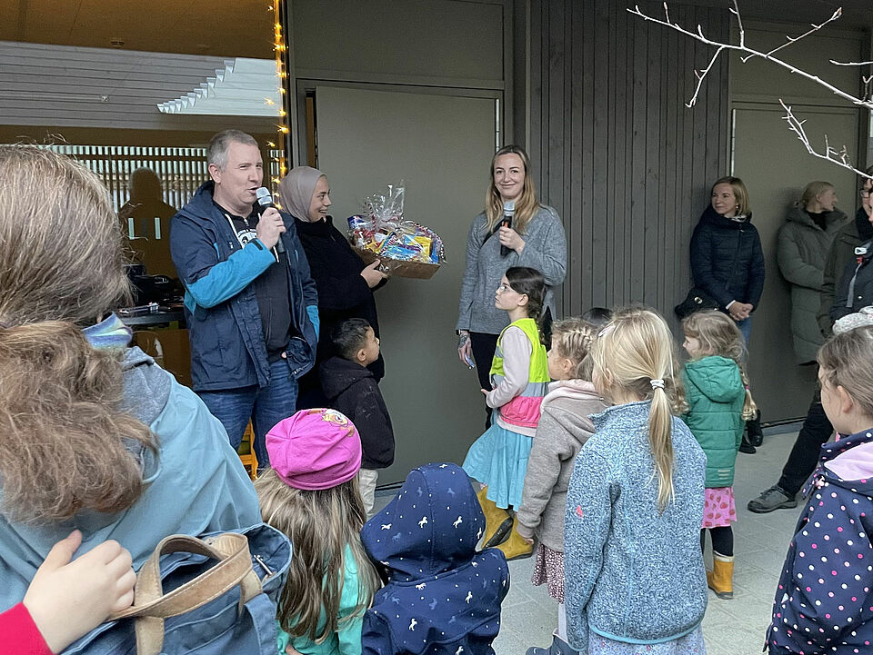Herbstfest Drei Personen stehen vor einem modernen Gebäude, eine Person hat ein Mikrofon in der Hand, eine Person überreicht einen Präsentkorb. Vor ihnen stehen mehrere Kinder und im Hintergrund sind weitere Personen zu sehen.