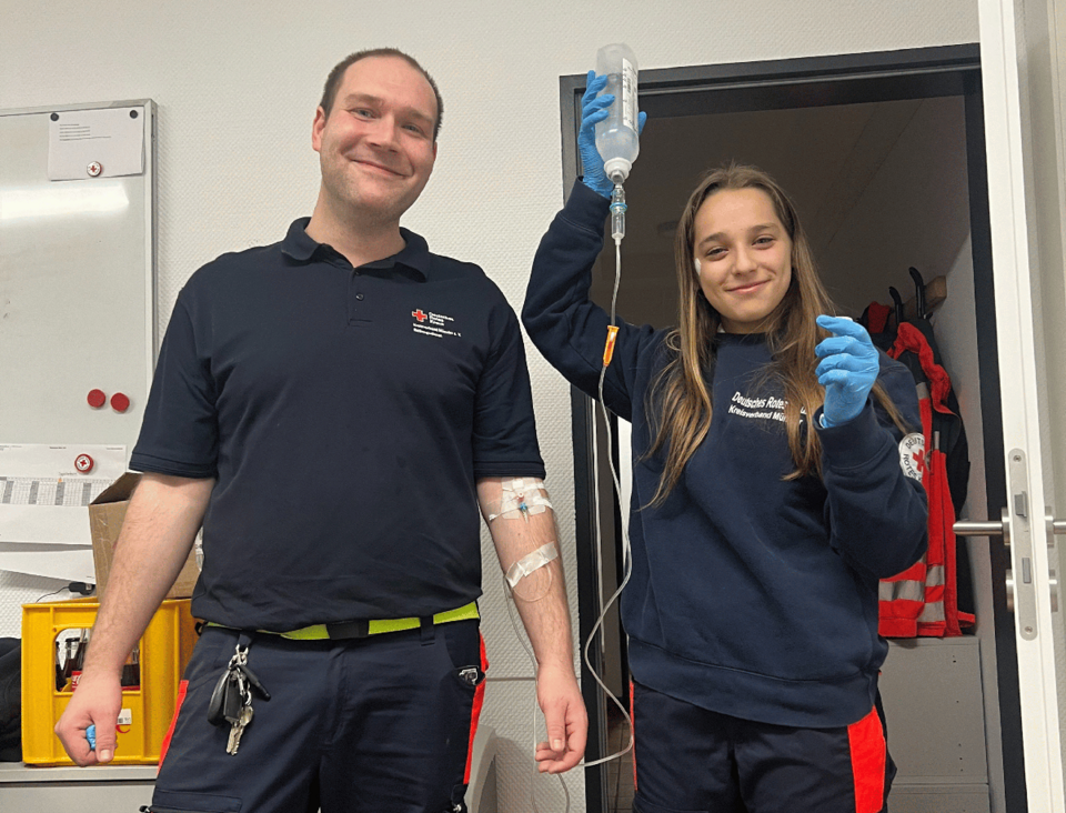 Zugang Ein Mann und ein Mädchen in Einsatzkleidung, er hat einen Zugang angelegt, sie hält die Flüssigskeitsflasche stolz in die Höhe