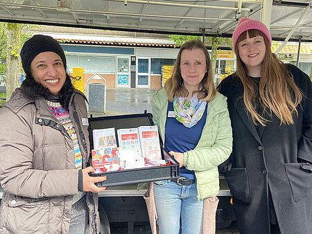 Drei Frauen halten einen Bauchladen mit Flyern in die Kamera