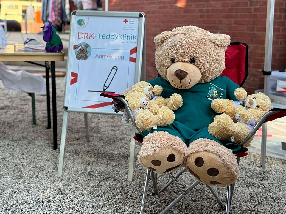 Ein großer Teddy trägt ein JRK-Shirt und sitzt in einem Campingstuhl und hat weitere kleinere Teddys im Arm. Im Hintergrund ist ein DRK-Teddyklinik Anmeldung Schild zu sehen