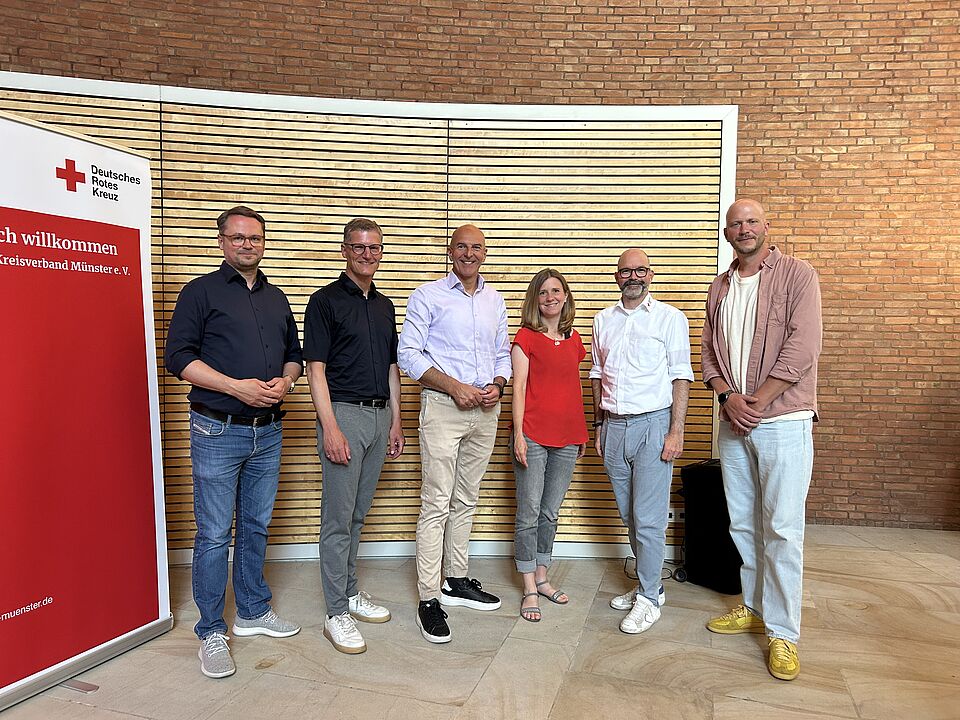 Sechs Personen stehen für ein Gruppenfoto vor einer Holz- und Backsteinwand. Links ist ein Banner des Deutschen Roten Kreuzes mit Willkommensgruß zu sehen