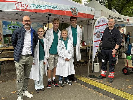 Gruppenbild vor einem weißem Pavilion mit der Aufschrift "DRK-Teddyklinik". Vier Kinder und Jugendliche in JRK-Oberteilen und weißem Kittel in der Mitte, recht eine Person in Einsatzkleidung, links eine Person in DRK-Jacke
