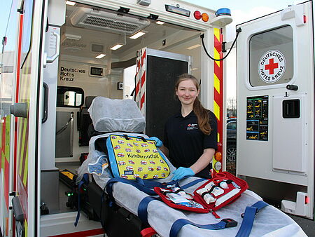 Eine junge Frau steht an der offenen Hecktür eines Rettungswagens mit ausgefahrener Trage