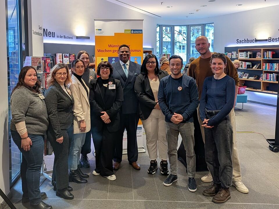 Eine Gruppe von Menschen steht vor einem "Wochen gegen Rassismus" Banner, im Hintergrund sind Bücherregale zu sehen.