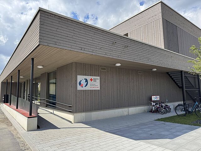 Ein modernes Gebäude mit einem Logo der Kita und einem Rotkreuzlogo und der Aufschrift "DRK Münster Sozialwerk gGmbH Kita Hand in Hand" Rechts im Bild befinden sich Fahrradständer.