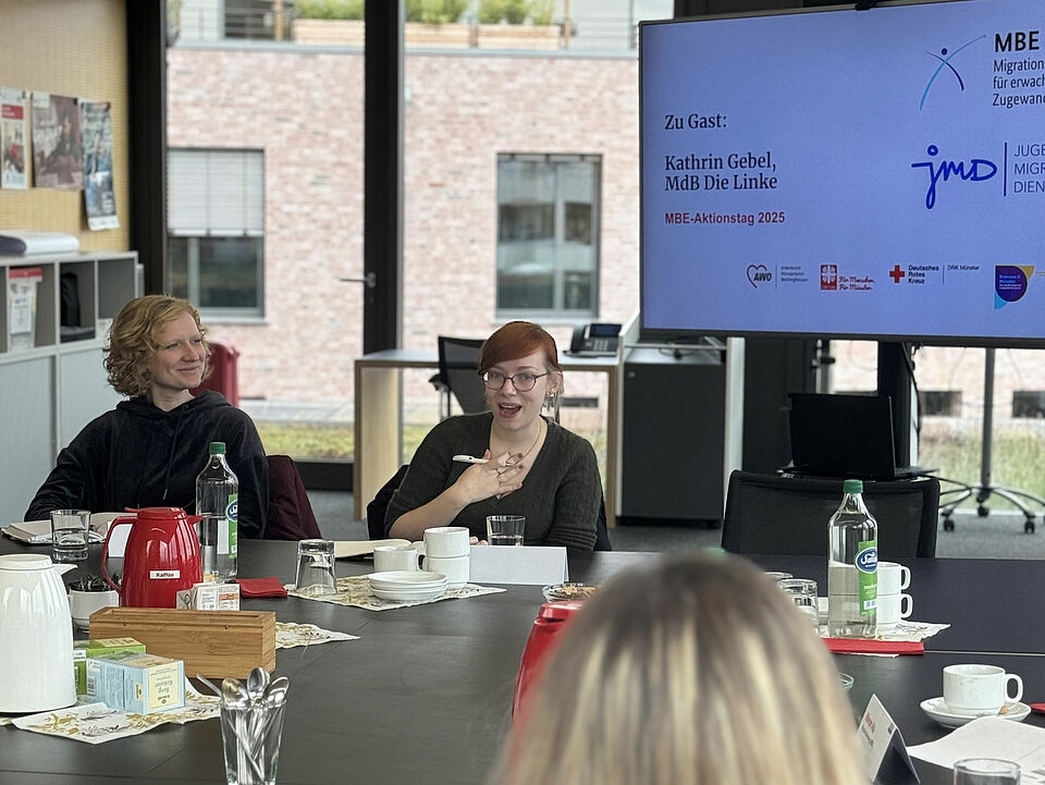 Gebel Zwei Frauen sitzen an einem Tisch mit Getränken und Snacks. Eine Person spricht und hält die Hand auf der Brust.