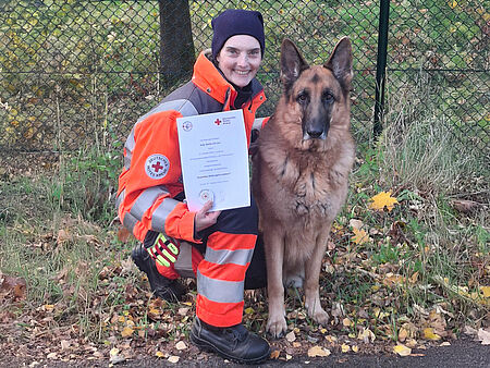 Eine DRK-Einsatzkraft kniet neben einem Hund mit einer Urkunde in der Hand.