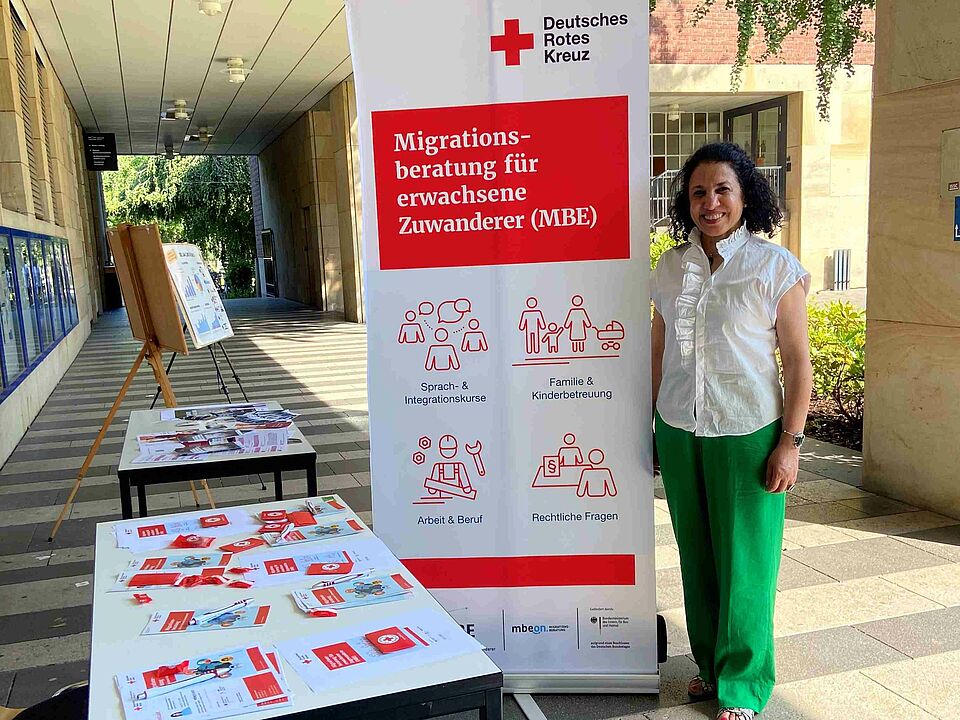 MBE bei Law Clinic Münster Eine Frau steht neben einem Tisch mit Infomaterial und einem Roll-Up zur Migrationsberatung.