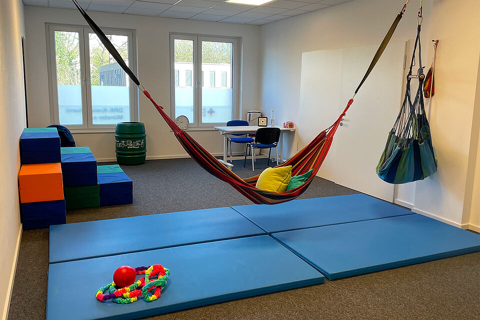 Ein Raum mit 2 Fenstern, blauen Sportmatten, 2 Hängematten und bunten Schaumstoffwürfeln