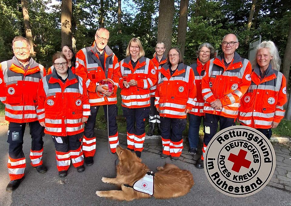 Lupus Eine Gruppe von Personen in DRK-Einsatzkleidung, im Vordergrund liegt ein Hund. Rechts auf dem Bild ist eine Darstellung der DRK-Rettungshund im Ruhestand Plakette zu sehen