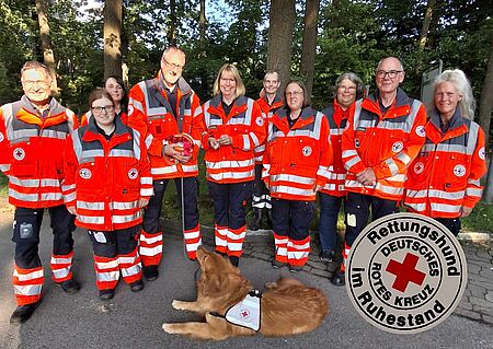 Eine Gruppe von Personen in DRK-Einsatzkleidung, im Vordergrund liegt ein Hund. Rechts auf dem Bild ist eine Darstellung der DRK-Rettungshund im Ruhestand Plakette zu sehen