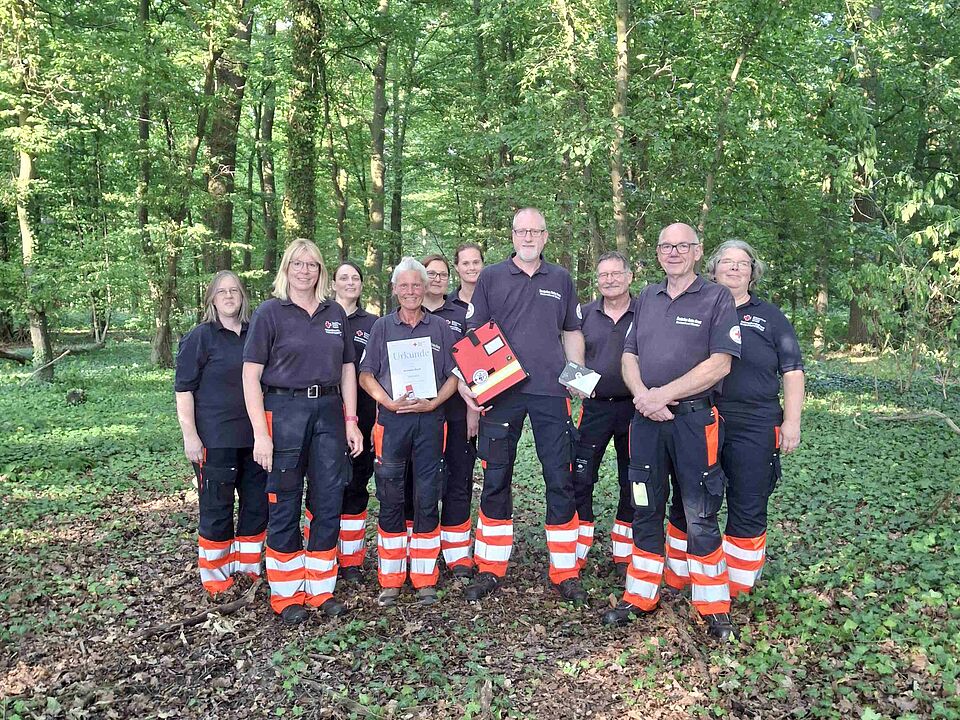 Maintrailing-Prüferin Gruppe von Personen in Einsatzkleidung in einem Wald. Frau in der Mitte hält eine Urkunde in der Hand. Ein Mann eine Mappe und eine Box.