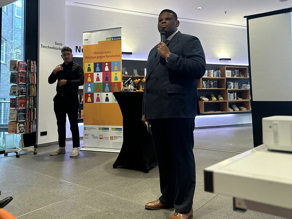Mann spricht in ein Mikrofon. Im Hintergrund steht ein Stehtisch und ein Banner mit dem Logo der Wochen gegen Rassismus, dahinter sind Bücherregale zu sehen.